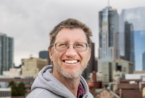 Dr. John Carter smiling with the Denver skyline behind him En español: Dr. John Carter sonriendo con el horizonte de Denver detrás de él