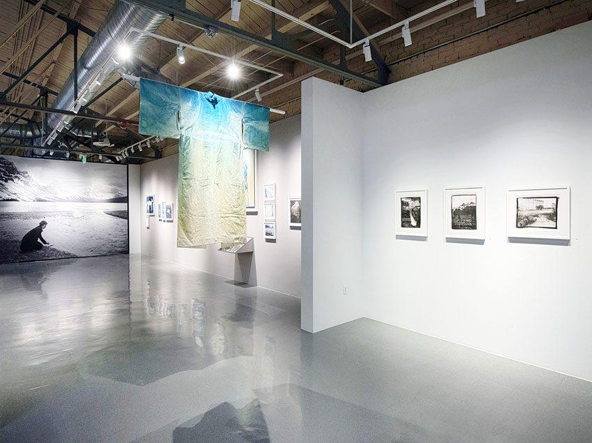 Photograph of gallery space at CVA. On the left wall there are framed photographic prints. Hanging in the middle of the gallery is a blue and yellow yukata. At the end of the hallway is a large black and white mural of a woman kneeling at the edge of a lake, where there are snowy mountains in the background.